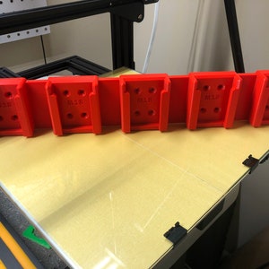 May include: A red 3D printed wall mount with five slots for holding objects. Each slot is labeled "M18" and has multiple holes for screws.
