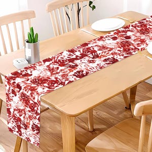 May include: A rectangular table runner featuring a white background with a repeating pattern of red handprints. The design covers the entire length of the fabric, which is draped across a light-colored wooden dining table with chairs.