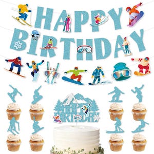 Op de afbeelding: Een feestelijke verjaardagstaart met cupcakes en decoraties. De taart is wit met een blauwe glitter berg-thema topper. De banner leest "HAPPY BIRTHDAY" met skiër- en snowboarder uitsparingen.
