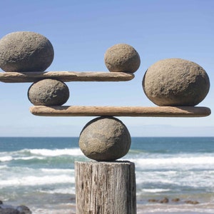 Peut inclure: Une sculpture de pierres équilibrées avec cinq pierres lisses de couleur grise empilées sur deux poutres en bois. La sculpture est placée sur fond d'océan bleu avec des vagues blanches.