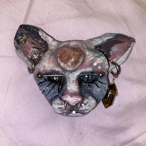 Op de afbeelding: Handgeschilderde kattengezichtsculptuur met een hartvormige steen op het voorhoofd. De sculptuur heeft blauwe, roze en witte verf, met metalen piercings en een bungelende kristallen oorbel. Het gezicht van de kat heeft een serene uitdrukking.