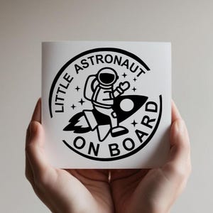 May include: A square white decal featuring a black circular graphic of an astronaut riding a rocket ship. The text LITTLE ASTRONAUT ON BOARD is printed in a bold black sans-serif font along the circular border, accented by small star icons.