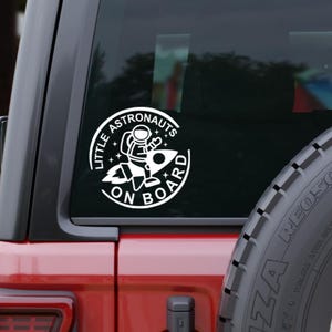 May include: A circular white vinyl decal applied to a vehicle window featuring a graphic of an astronaut riding a rocket ship. The text "LITTLE ASTRONAUTS ON BOARD" is printed in a bold, sans-serif font around the circular border with stars.