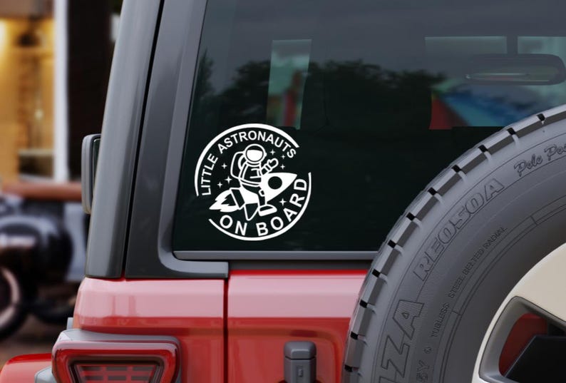 May include: A circular white vinyl decal applied to a vehicle window featuring a graphic of an astronaut riding a rocket ship. The text "LITTLE ASTRONAUTS ON BOARD" is printed in a bold, sans-serif font around the circular border with stars.