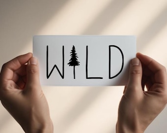 Puede incluir: Una pegatina rectangular blanca con la palabra WILD en letras mayúsculas negras sin remates. La letra I está sustituida por la silueta negra de un árbol de coníferas. El diseño es limpio, minimalista y está centrado en el fondo blanco.