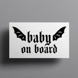 Puede incluir: Una pegatina rectangular blanca con texto negro que dice baby on board en una fuente de estilo gótico. El diseño incluye dos alas de murciélago negras que se extienden desde los lados de la palabra baby, creando un elemento gráfico simétrico.