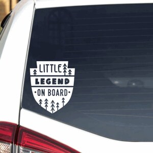 May include: White car window sticker with black text that reads "Little Legend On Board" surrounded by a design of small pine trees.