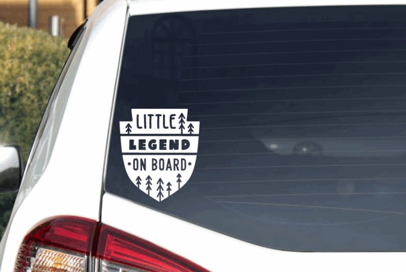 May include: White car window sticker with black text that reads "Little Legend On Board" surrounded by a design of small pine trees.