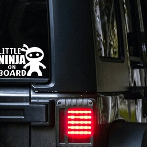 Puede incluir: Calcoman&iacute;a de vinilo blanco con el texto "Little Ninja On Board" y un ninja de dibujos animados con una m&aacute;scara negra. La calcoman&iacute;a est&aacute; en la ventana trasera de un Jeep negro.