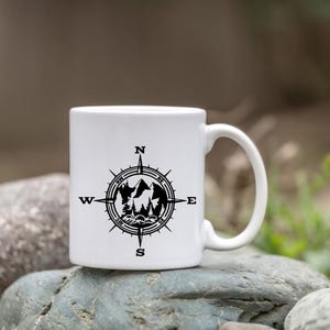 Puede incluir: Una taza de cer&aacute;mica blanca con un dise&ntilde;o gr&aacute;fico negro de una rosa de los vientos. El centro de la br&uacute;jula contiene una ilustraci&oacute;n en silueta de picos de monta&ntilde;as, &aacute;rboles de hoja perenne y olas. Los puntos cardinales N, E, S, O est&aacute;n impresos.