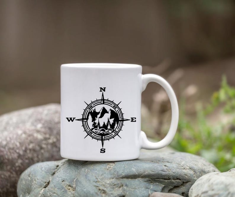 Puede incluir: Una taza de cer&aacute;mica blanca con un dise&ntilde;o gr&aacute;fico negro de una rosa de los vientos. El centro de la br&uacute;jula contiene una ilustraci&oacute;n en silueta de picos de monta&ntilde;as, &aacute;rboles de hoja perenne y olas. Los puntos cardinales N, E, S, O est&aacute;n impresos.