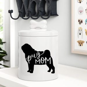 May include: White ceramic jar with a bone-shaped lid. The jar has a black silhouette of a pug with the text "pug mom" printed on it.