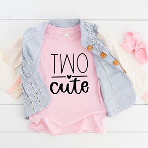 Puede incluir: Camiseta rosa para niño pequeño con el texto "TWO cute" en letras negras con un pequeño corazón sobre la palabra "cute". La camiseta se muestra sobre una superficie blanca con una chaqueta vaquera y un lazo rosa.