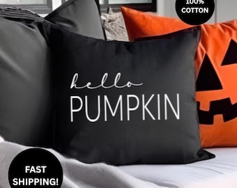 May include: A square black throw pillow featuring the white text hello PUMPKIN in a mix of script and sans-serif fonts. The pillow is made of 100% cotton and is positioned next to an orange pillow with a black jack-o'-lantern face design.