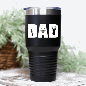 Pu&ograve; includere: Un bicchiere in acciaio inossidabile nero con un design bianco che dice "DAD" con un fucile, un bersaglio e una silhouette di cervo.