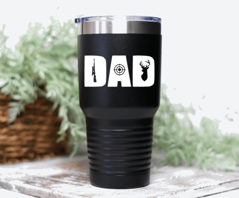 Pu&ograve; includere: Un bicchiere in acciaio inossidabile nero con un design bianco che dice "DAD" con un fucile, un bersaglio e una silhouette di cervo.