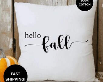 May include: A square white throw pillow featuring the black text hello fall in a mix of serif and script fonts. A black circular badge in the top right corner reads 100% COTTON, and a black circular badge in the bottom left reads FAST SHIPPING.
