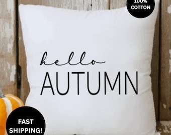May include: A square white throw pillow featuring the black text hello AUTUMN centered on the front. A black circular badge in the top right corner displays the text 100% COTTON. The pillow is positioned in front of a light wood background.