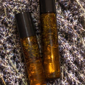 Può includere: Due flaconi roll-on in vetro ambrato con tappi neri, contenenti ciascuno un liquido dorato infuso con boccioli di lavanda essiccati. I flaconi sono posizionati verticalmente, mettendo in risalto il vetro trasparente e il contenuto botanico.
