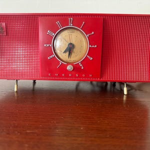 Puede incluir: Un radio despertador vintage Emerson rojo con una esfera blanca y n&uacute;meros negros. La radio tiene un panel frontal texturizado y un peque&ntilde;o logotipo en el lado izquierdo. El radio despertador est&aacute; sobre una superficie de madera.