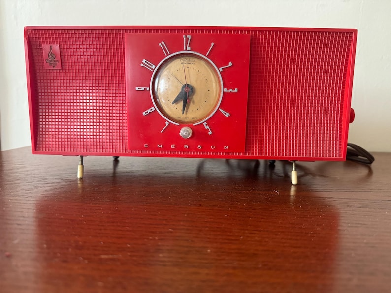 Puede incluir: Un radio despertador vintage Emerson rojo con una esfera blanca y n&uacute;meros negros. La radio tiene un panel frontal texturizado y un peque&ntilde;o logotipo en el lado izquierdo. El radio despertador est&aacute; sobre una superficie de madera.