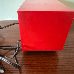 Puede incluir: Una caja rectangular de color rojo brillante con un cable negro conectado a ella. La caja est&aacute; sobre una superficie de madera.