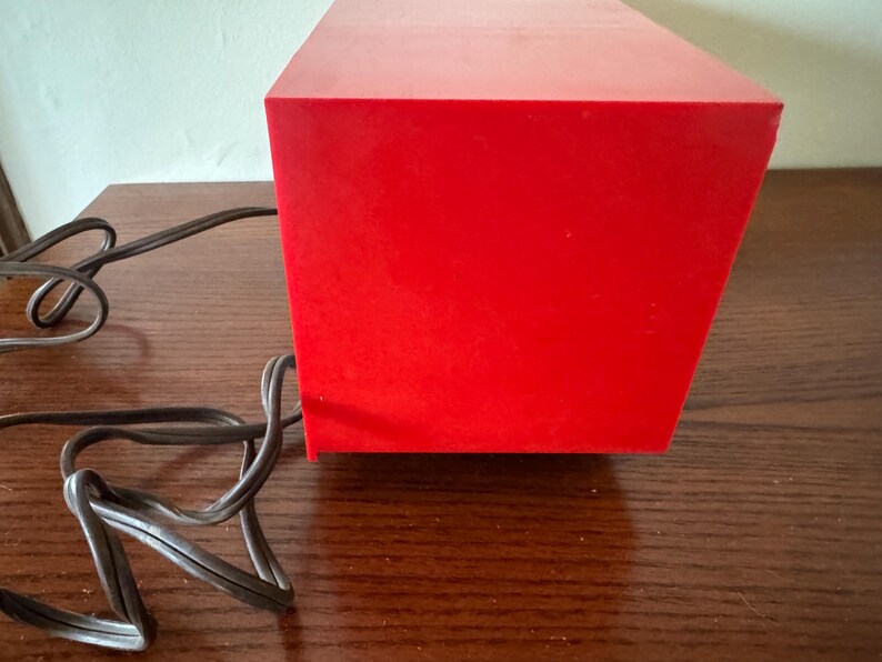 Puede incluir: Una caja rectangular de color rojo brillante con un cable negro conectado a ella. La caja est&aacute; sobre una superficie de madera.