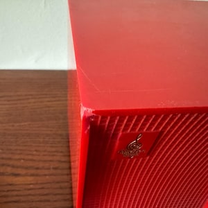 Puede incluir: Un altavoz rojo con una superficie texturizada y un logotipo dorado con una clave de sol. El altavoz tiene una esquina ligeramente da&ntilde;ada.