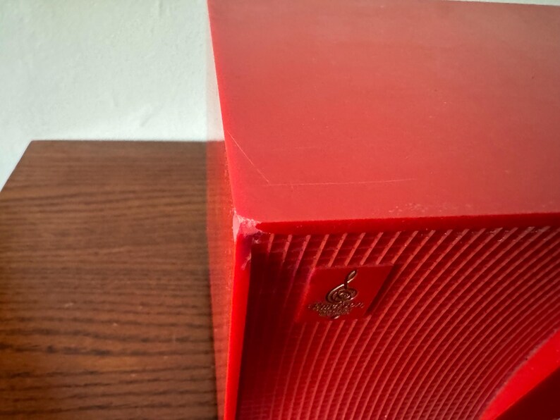 Puede incluir: Un altavoz rojo con una superficie texturizada y un logotipo dorado con una clave de sol. El altavoz tiene una esquina ligeramente da&ntilde;ada.