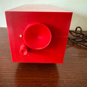 Puede incluir: Una radio vintage roja con un altavoz redondo grande y una perilla m&aacute;s peque&ntilde;a. La radio est&aacute; sobre una superficie de madera y tiene un cable negro.