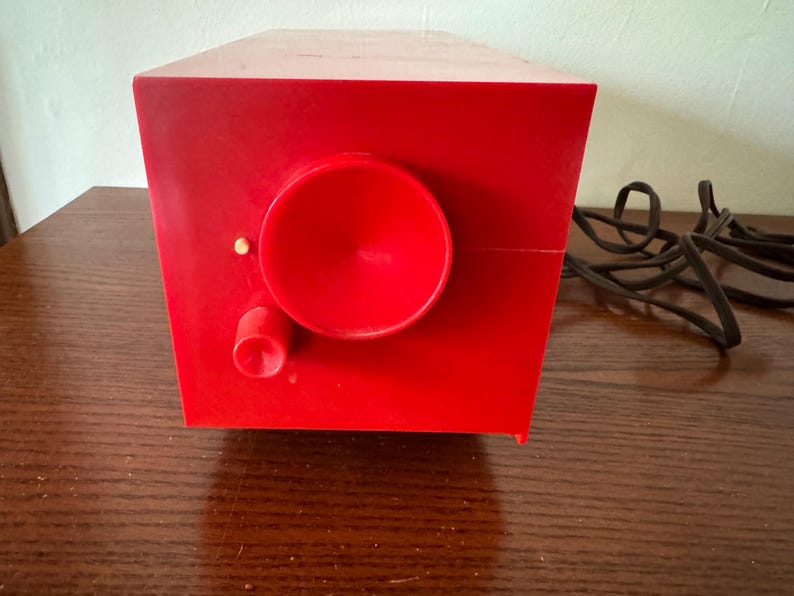 Puede incluir: Una radio vintage roja con un altavoz redondo grande y una perilla m&aacute;s peque&ntilde;a. La radio est&aacute; sobre una superficie de madera y tiene un cable negro.
