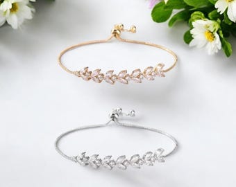 May include: Two adjustable bracelets. One is gold-toned, the other silver-toned. Both feature a delicate leaf design with clear, sparkling accents. The bracelets are displayed on a white surface, with white flowers and green leaves in the background.