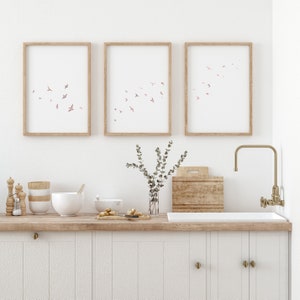 Puede incluir: Tres impresiones enmarcadas con un dise&ntilde;o de acuarela de p&aacute;jaros rosas volando sobre un fondo blanco. Las impresiones est&aacute;n colgadas en una pared blanca sobre una encimera de cocina con una encimera de madera y gabinetes blancos. Hay un jarr&oacute;n con vegetaci&oacute;n, una tabla de cortar de madera y un fregadero con un grifo dorado.
