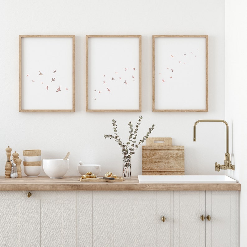 Puede incluir: Tres impresiones enmarcadas con un dise&ntilde;o de acuarela de p&aacute;jaros rosas volando sobre un fondo blanco. Las impresiones est&aacute;n colgadas en una pared blanca sobre una encimera de cocina con una encimera de madera y gabinetes blancos. Hay un jarr&oacute;n con vegetaci&oacute;n, una tabla de cortar de madera y un fregadero con un grifo dorado.