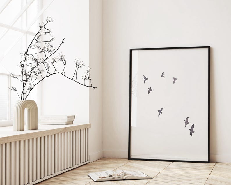 May include: A framed art print featuring a minimalist design of several birds in flight against a white background. The artwork is in a black frame and is displayed in a bright, airy room with a window and decorative vase.