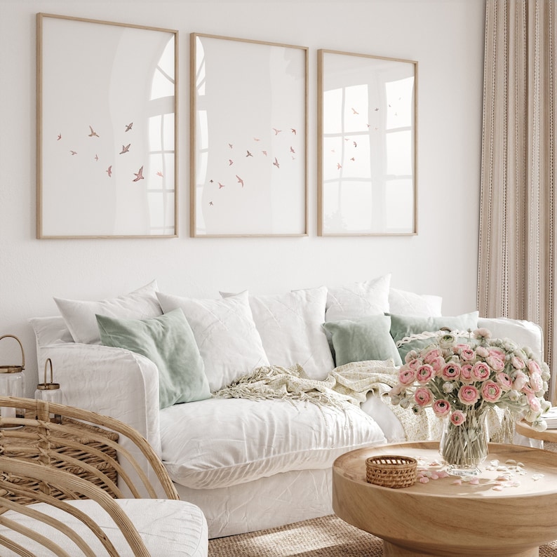 May include: A white couch with light green and white pillows, a white throw blanket, and a vase of pink flowers on a wooden coffee table. Three framed prints with a flock of birds in flight are hanging on the wall behind the couch.