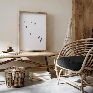 May include: A framed print with a minimalist design of flying birds hangs above a bamboo bench. A woven basket and a curved rattan chair with a black cushion are in the foreground. The room has a neutral color palette.