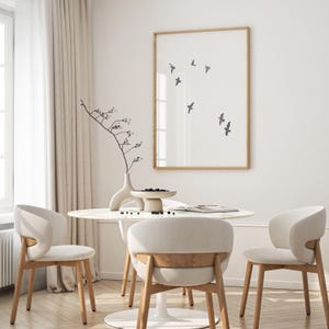May include: A dining room scene featuring a round marble table with three cream-colored chairs. A framed print with flying birds hangs on the wall. A vase with a decorative branch and a small bowl of berries complete the decor.