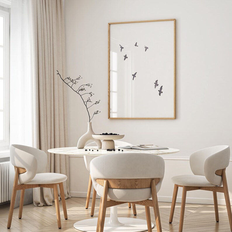 May include: A dining room scene featuring a round marble table with three cream-colored chairs. A framed print with flying birds hangs on the wall. A vase with a decorative branch and a small bowl of berries complete the decor.
