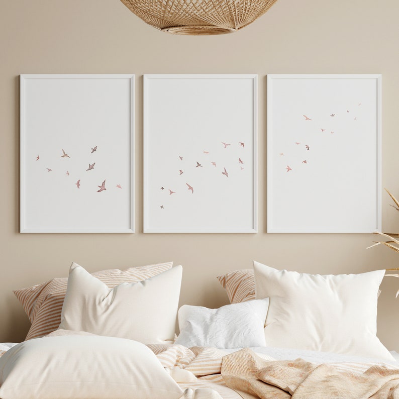 Puede incluir: Tres impresiones enmarcadas blancas con un dise&ntilde;o minimalista de p&aacute;jaros rosas volando en un degradado. Las impresiones est&aacute;n colgadas en una pared beige claro sobre una cama con ropa de cama a rayas blancas y marrones.