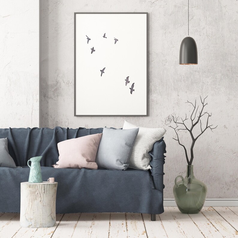 May include: A framed art print featuring a minimalist design of several birds in flight against a white background. The artwork hangs above a blue sofa with decorative pillows. A vase with branches and a hanging lamp complete the room's decor.
