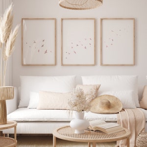 Puede incluir: Tres impresiones enmarcadas con un dise&ntilde;o minimalista que presenta una bandada de p&aacute;jaros rosas volando sobre un fondo blanco. Las impresiones est&aacute;n colgadas en una pared blanca sobre un sof&aacute; blanco con almohadas beige y una manta de tejido.
