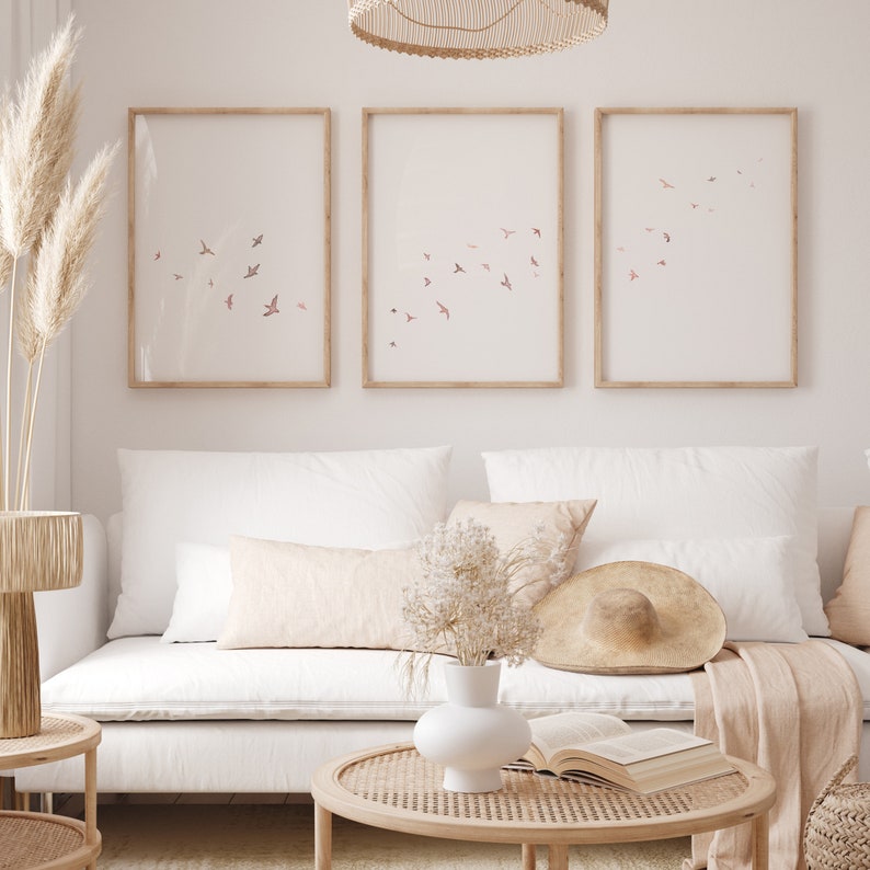 Puede incluir: Tres impresiones enmarcadas con un dise&ntilde;o minimalista que presenta una bandada de p&aacute;jaros rosas volando sobre un fondo blanco. Las impresiones est&aacute;n colgadas en una pared blanca sobre un sof&aacute; blanco con almohadas beige y una manta de tejido.