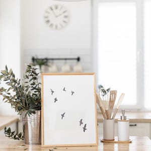 May include: A framed print with a minimalist design of flying birds. The artwork is in a light wood frame and is displayed on a wooden surface. A metal bucket with greenery, kitchen utensils, and a soap dispenser are also visible.