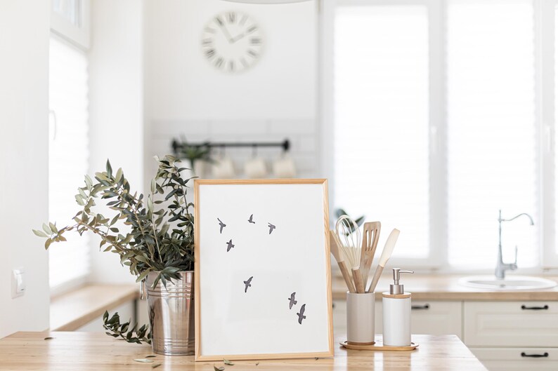 May include: A framed print with a minimalist design of flying birds. The artwork is in a light wood frame and is displayed on a wooden surface. A metal bucket with greenery, kitchen utensils, and a soap dispenser are also visible.