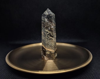 Könnte beinhalten: Eine klare Quarzkristallspitze mit schwarzen und grünen Einschlüssen, die auf einem goldfarbenen Tablett steht.