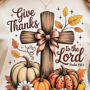 May include: Cream-colored t-shirt featuring a graphic design with the words "Give Thanks to the Lord" and "Psalm 136:1". The design includes a wooden cross with a bow, pumpkins, leaves, and daisies.