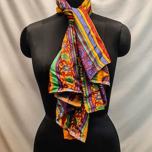 May include: A colorful patterned scarf with yellow, red, green, and blue stripes and floral designs. The scarf is tied around a black mannequin's neck.