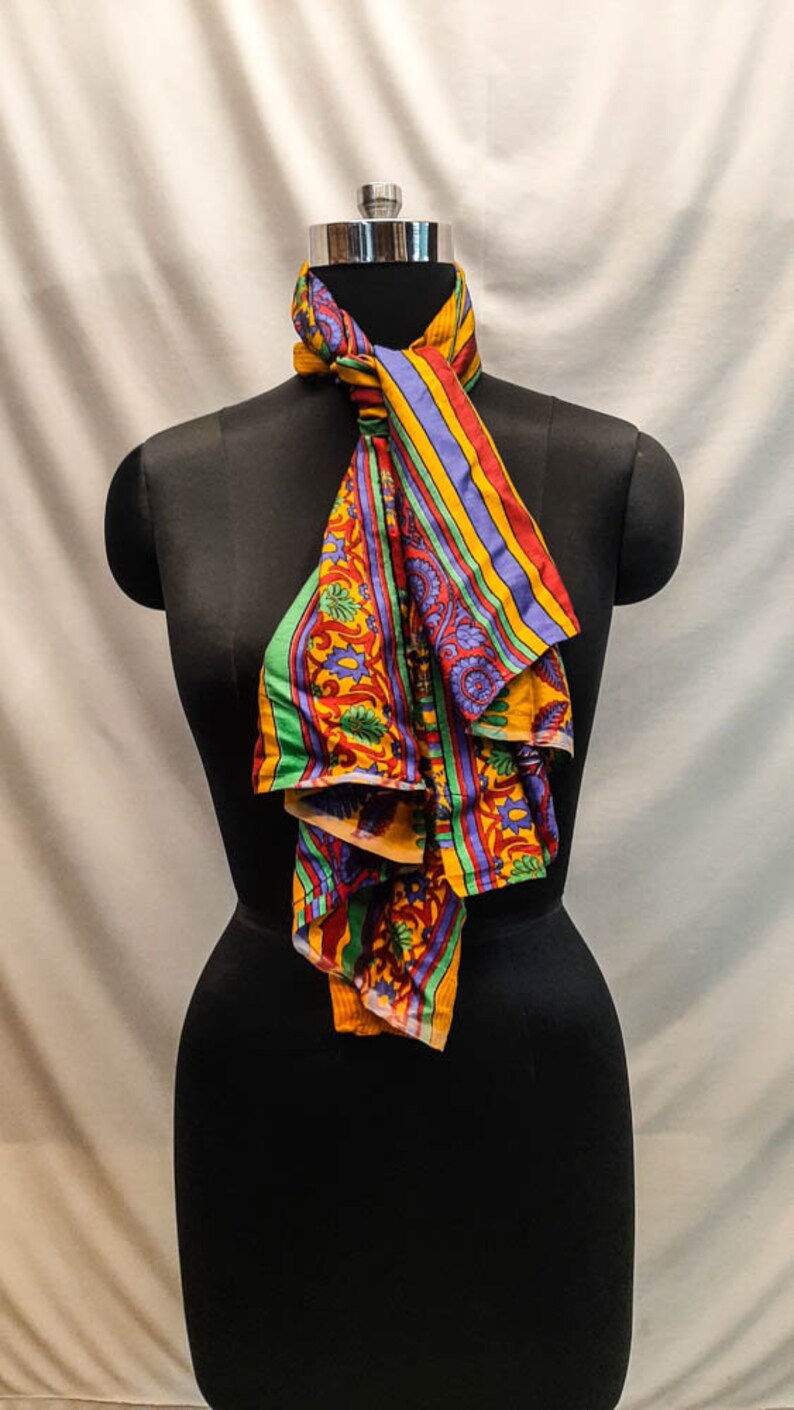 May include: A colorful patterned scarf with yellow, red, green, and blue stripes and floral designs. The scarf is tied around a black mannequin's neck.