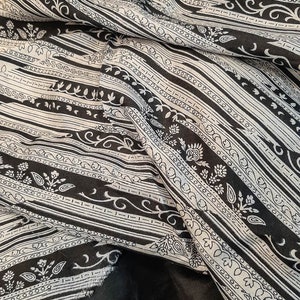May include: A black and white patterned fabric with a repeating design of stripes and floral motifs. The fabric is folded and draped, showing the texture and detail of the pattern.